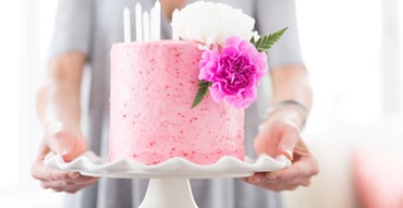 A closeup of arms presenting a pink cake with flowers on it.